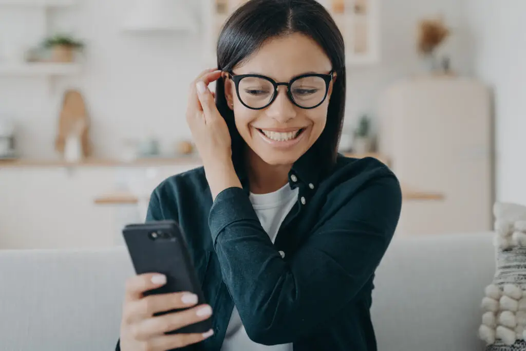 Small business marketing in Kamloops example photo shows a smiling woman holding smartphone reading message chatting in social networks, sitting on couch at home. Portrait of excited girl wearing glasses received good news, got promotion at work.